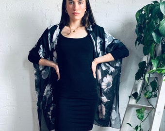 May include: A woman wearing a black dress with a floral print sheer overlay. She is standing in front of a white ladder with plants.