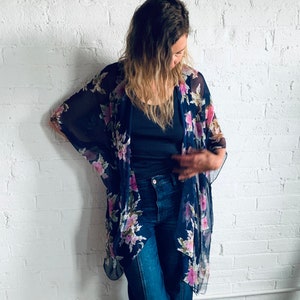 May include: A woman wearing a navy blue floral kimono with a black vest top and dark blue wide-leg jeans. She is standing in front of a white brick wall and wearing brown sandals.