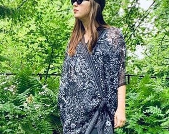 May include: A woman wearing a black and white patterned kimono-style top with black pants and brown sandals. She is standing in a garden with green foliage.