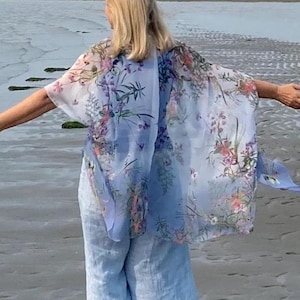 May include: A woman wearing a light blue floral patterned kimono with long sleeves and matching trousers stands on a sandy beach with her arms outstretched. The beach is wet and the water is receding in the background.