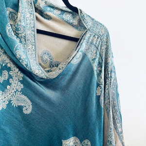 May include: A teal blue and cream paisley patterned shawl with a cowl neck. The shawl is hanging on a black hanger.
