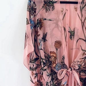 May include: A light pink kimono-style cover-up with a floral and hummingbird print. The open-front garment features a tie closure and flowing sleeves. The design includes various flowers and foliage in shades of brown, blue, and green.