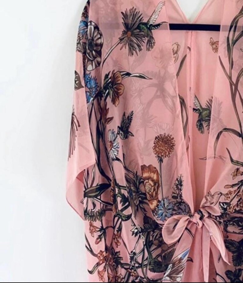 May include: A light pink kimono-style cover-up with a floral and hummingbird print. The open-front garment features a tie closure and flowing sleeves. The design includes various flowers and foliage in shades of brown, blue, and green.