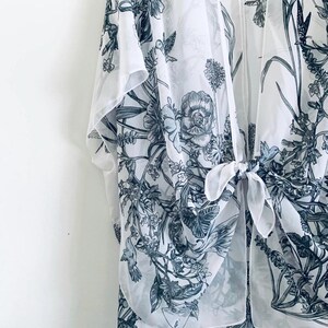 May include: A sheer white kimono-style cover-up with a botanical print in shades of gray and blue. The design features flowers, leaves, and birds. The garment has a tie closure at the front.