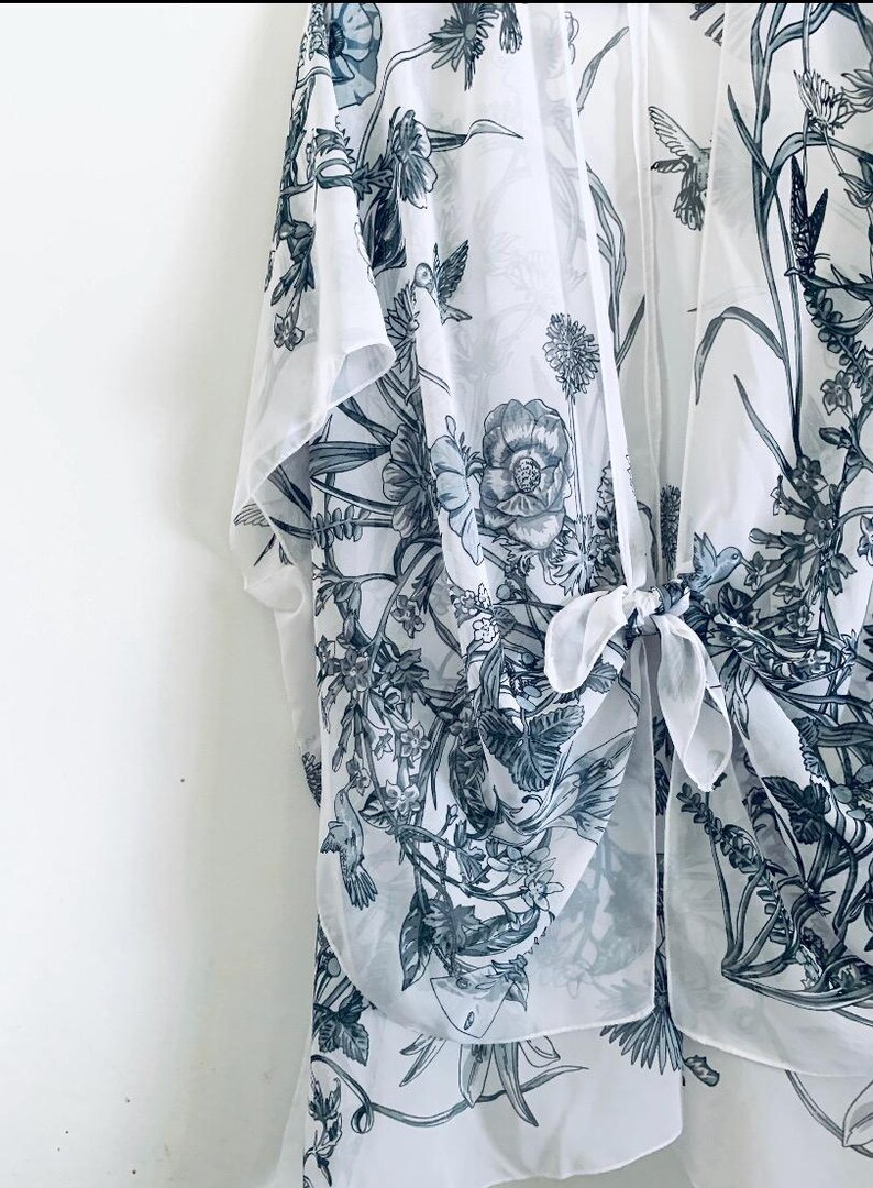 May include: A sheer white kimono-style cover-up with a botanical print in shades of gray and blue. The design features flowers, leaves, and birds. The garment has a tie closure at the front.