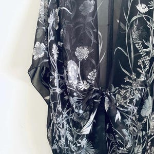 May include: Black sheer kimono with a floral pattern in shades of white and gray. The lightweight fabric drapes elegantly, featuring a tie closure at the waist. The design includes detailed botanical illustrations of flowers and foliage.