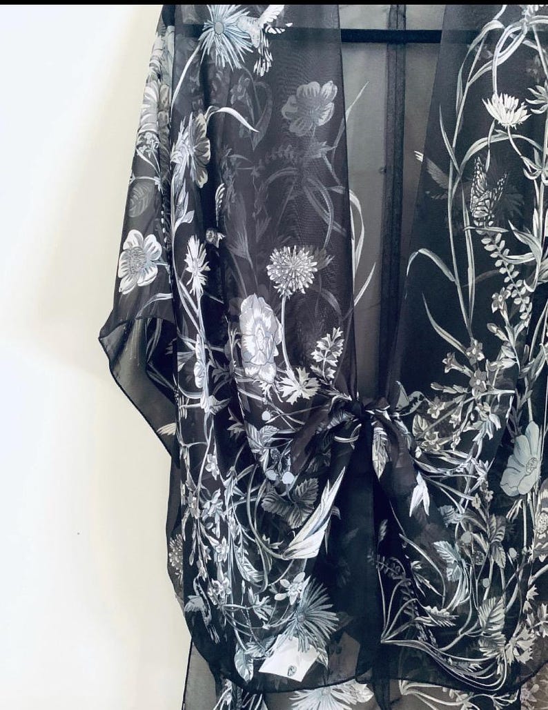 May include: Black sheer kimono with a floral pattern in shades of white and gray. The lightweight fabric drapes elegantly, featuring a tie closure at the waist. The design includes detailed botanical illustrations of flowers and foliage.