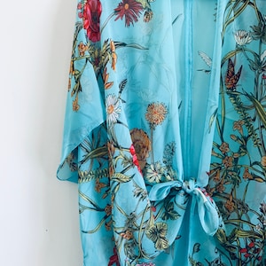 May include: A light blue kimono-style cover-up with a floral and butterfly print. The sheer fabric features red poppies, daisies, and other flowers. The cover-up is tied at the waist with a matching fabric tie.