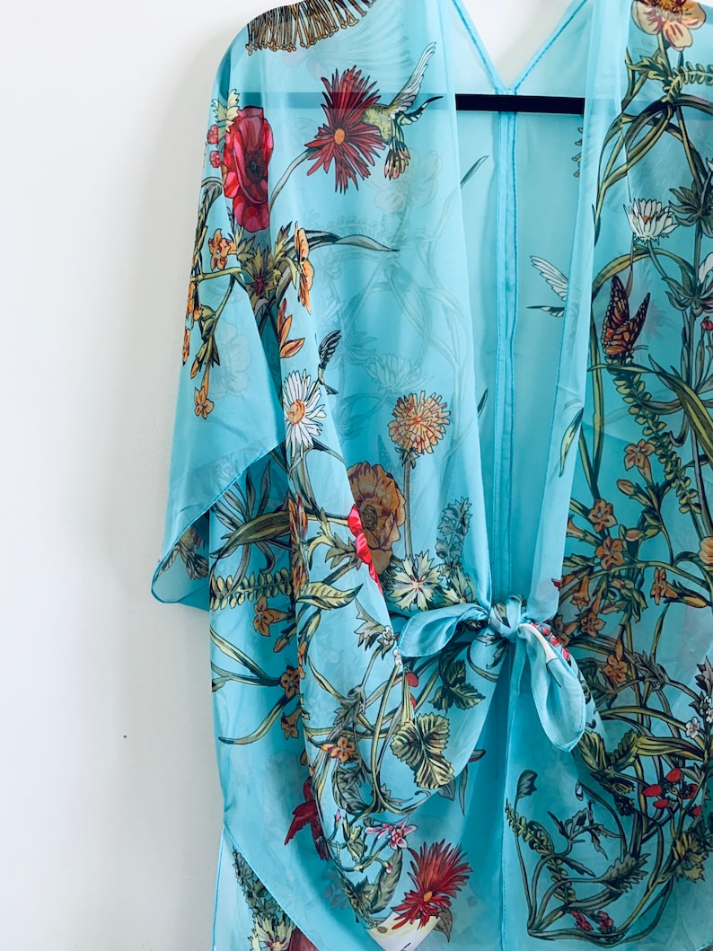 May include: A light blue kimono-style cover-up with a floral and butterfly print. The sheer fabric features red poppies, daisies, and other flowers. The cover-up is tied at the waist with a matching fabric tie.