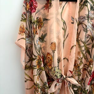 May include: A peach-colored kimono-style cover-up with a floral and hummingbird print. The sheer fabric features a tie closure at the waist and short sleeves. The design includes red, orange, yellow, and green floral elements.