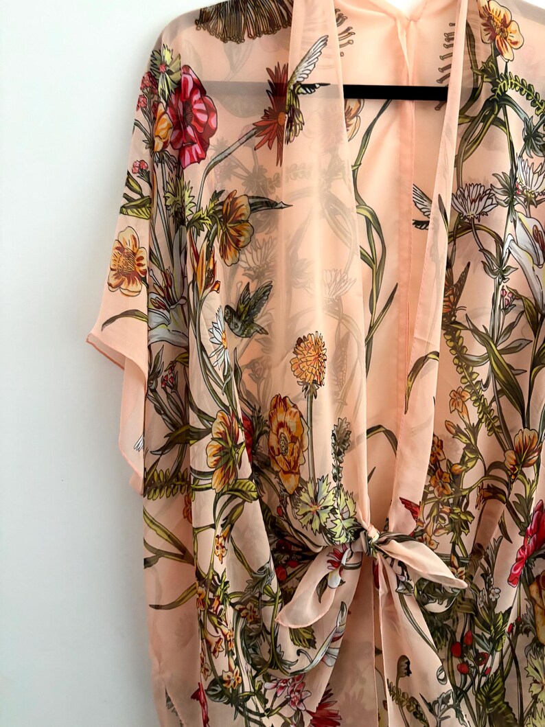 May include: A peach-colored kimono-style cover-up with a floral and hummingbird print. The sheer fabric features a tie closure at the waist and short sleeves. The design includes red, orange, yellow, and green floral elements.