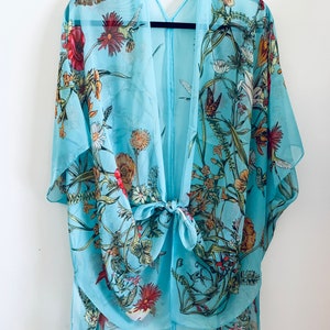 May include: A light blue kimono-style cover-up with a floral and butterfly print. The sheer fabric features red, yellow, and green flowers, along with butterflies and hummingbirds. The kimono has a tie closure at the waist and is hanging on a black hanger.