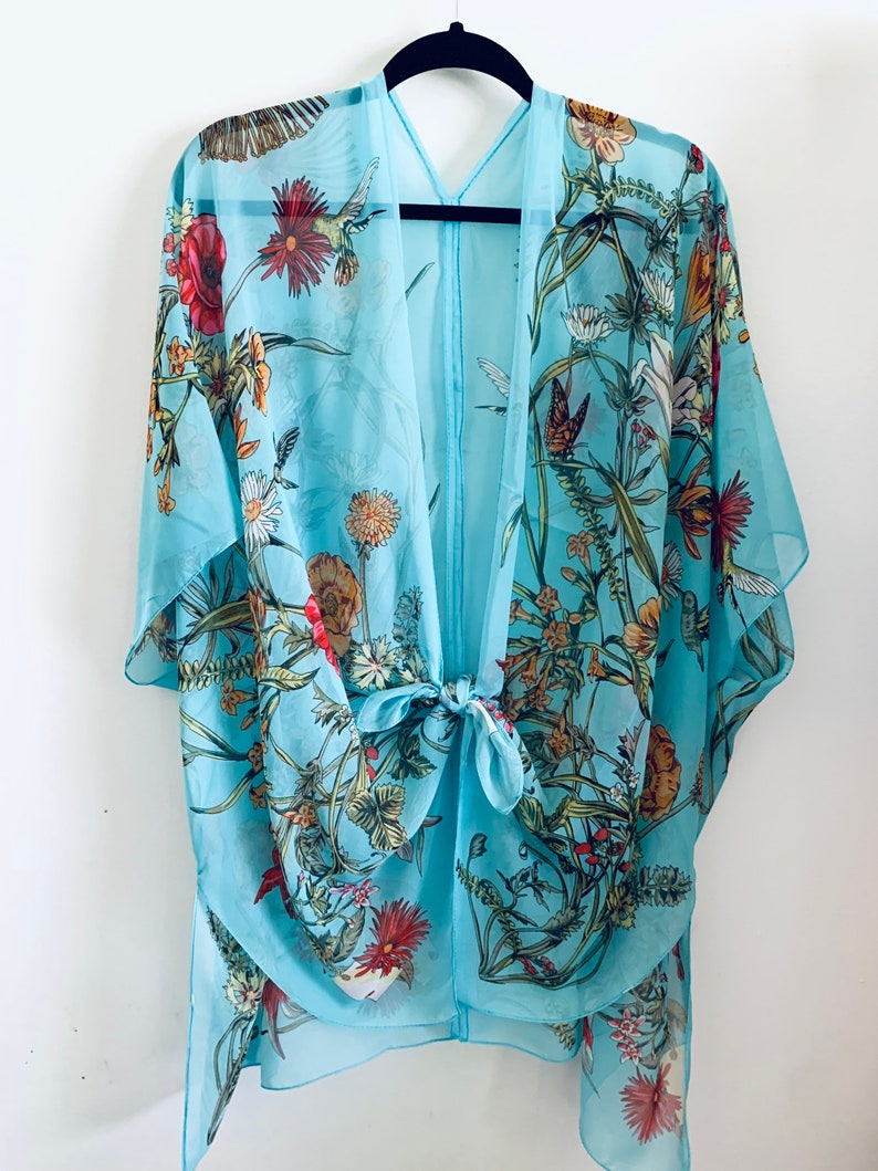 May include: A light blue kimono-style cover-up with a floral and butterfly print. The sheer fabric features red, yellow, and green flowers, along with butterflies and hummingbirds. The kimono has a tie closure at the waist and is hanging on a black hanger.