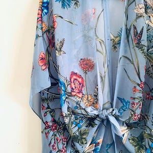 May include: A light blue floral kimono with a tie closure. The sheer fabric is adorned with colorful flowers, hummingbirds, and butterflies. The kimono is hanging on a black hanger.