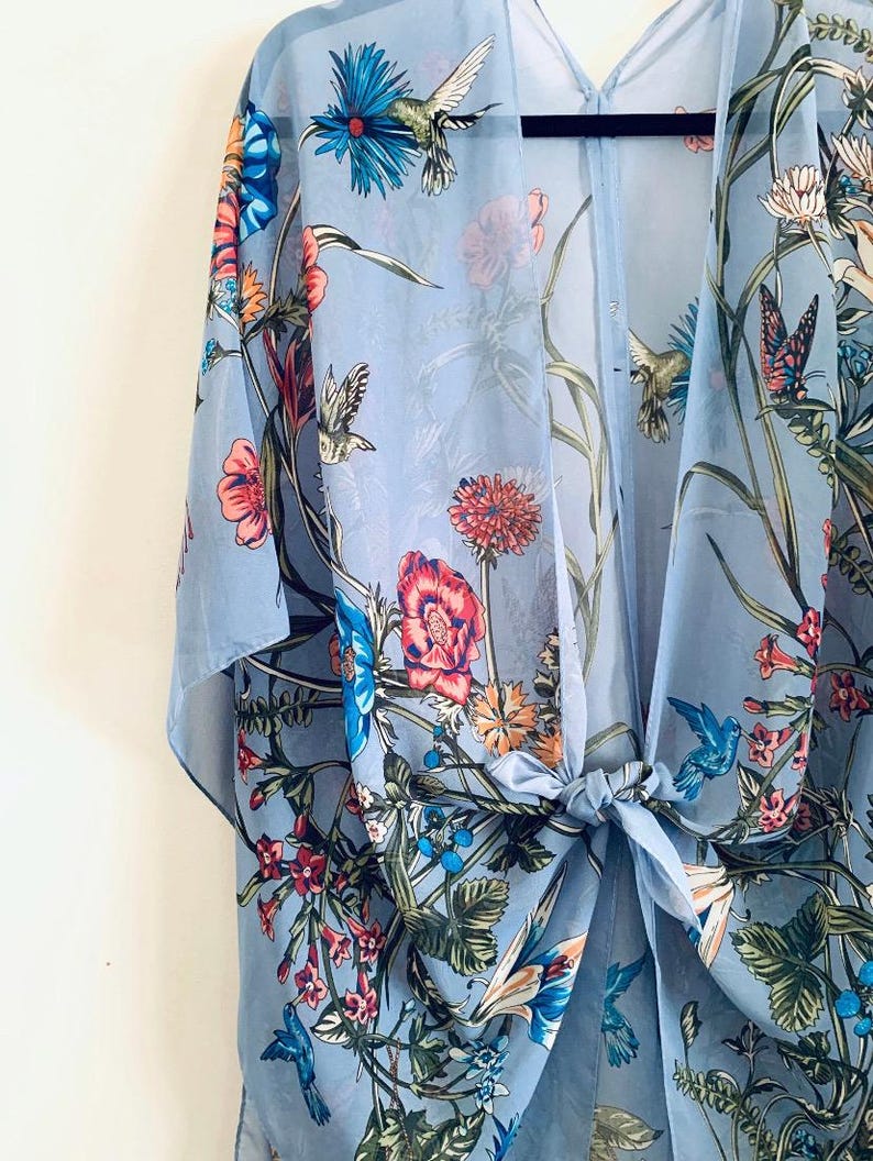 May include: A light blue floral kimono with a tie closure. The sheer fabric is adorned with colorful flowers, hummingbirds, and butterflies. The kimono is hanging on a black hanger.