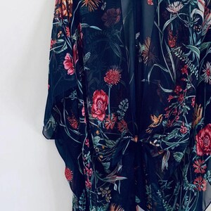May include: A dark navy blue kimono-style cover-up with a floral pattern. The sheer fabric features a vibrant design of red, pink, orange, and green flowers, leaves, and hummingbirds. The kimono is tied at the waist and hangs on a black hanger.