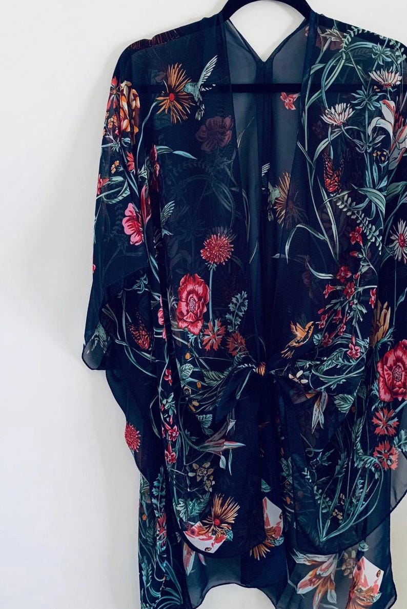 May include: A dark navy blue kimono-style cover-up with a floral pattern. The sheer fabric features a vibrant design of red, pink, orange, and green flowers, leaves, and hummingbirds. The kimono is tied at the waist and hangs on a black hanger.