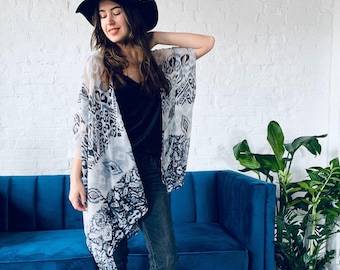 May include: A woman wearing a white and blue patterned kimono with a black hat and jeans. She is standing in front of a blue couch and a potted plant.