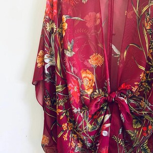 May include: A burgundy sheer kimono with a floral and hummingbird print. The open-front garment features a tie closure at the waist and is adorned with orange, red, and white flowers, along with green foliage and butterflies.