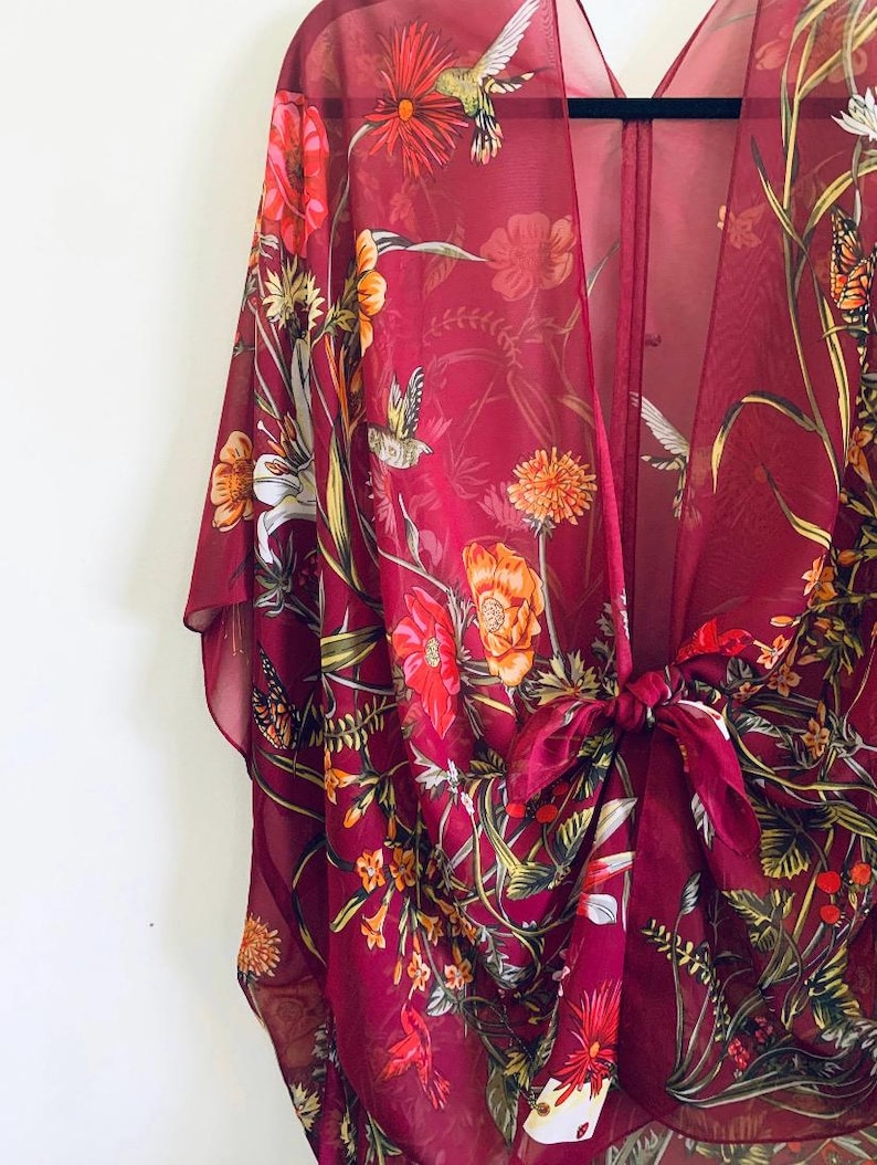 May include: A burgundy sheer kimono with a floral and hummingbird print. The open-front garment features a tie closure at the waist and is adorned with orange, red, and white flowers, along with green foliage and butterflies.