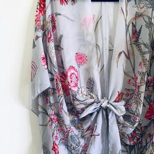 May include: A light gray kimono with a floral and hummingbird print. The kimono features pink and red flowers, green leaves, and a tie closure at the waist. The fabric is sheer and lightweight.