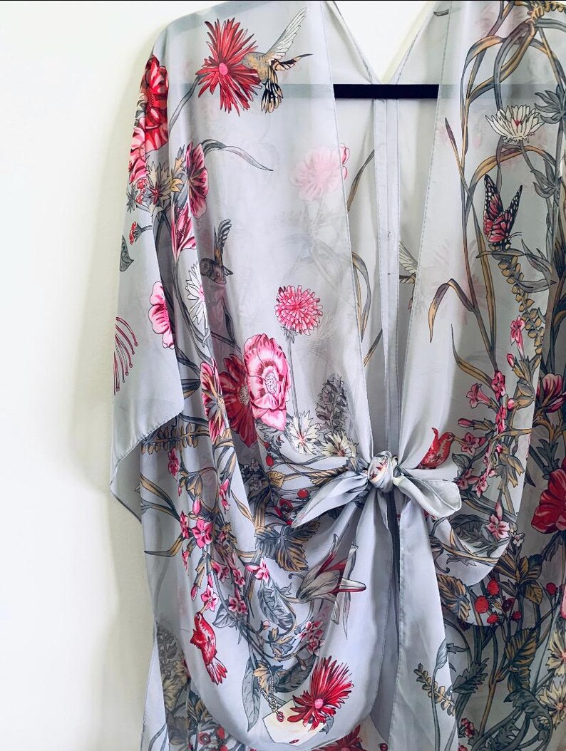 May include: A light gray kimono with a floral and hummingbird print. The kimono features pink and red flowers, green leaves, and a tie closure at the waist. The fabric is sheer and lightweight.