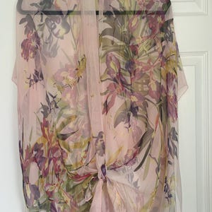 Peut inclure: Une écharpe rose pâle transparente ornée d'un motif floral aux tons violets, jaunes et verts. L'écharpe est suspendue à un cintre noir et nouée à l'avant. Le motif floral est visible sur toute la surface.