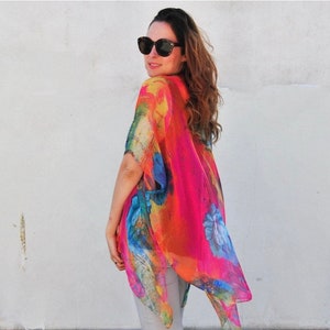 K&ouml;nnte beinhalten: Eine Frau tr&auml;gt einen farbenfrohen Kimono mit floralem Muster und rosa Grundfarbe. Der Kimono hat eine flie&szlig;ende, lockere Passform und ist aus einem leichten Stoff gefertigt. Die Frau tr&auml;gt eine Sonnenbrille und hat langes, braunes Haar.