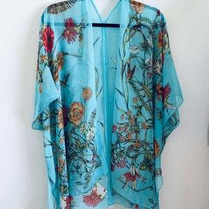 May include: A light blue kimono-style cover-up with a floral and bird print. The sheer fabric features red, orange, and white flowers, green leaves, and hummingbirds. The garment is hanging on a black hanger against a white background.