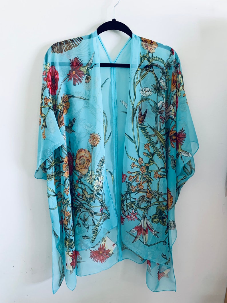 May include: A light blue kimono-style cover-up with a floral and bird print. The sheer fabric features red, orange, and white flowers, green leaves, and hummingbirds. The garment is hanging on a black hanger against a white background.