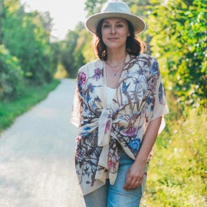 May include: A woman wearing a cream-colored kimono with a floral pattern of pink, blue, and brown. She is also wearing a light-colored hat, a white top, and blue jeans. The background is a path surrounded by green trees.