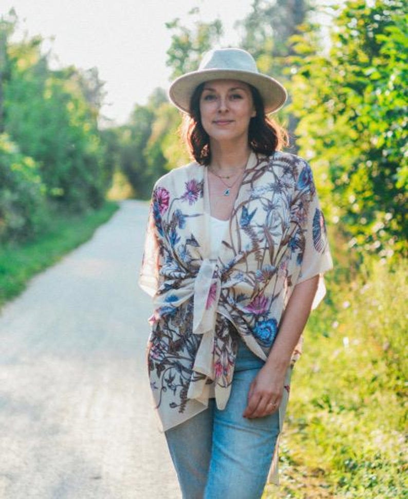 May include: A woman wearing a cream-colored kimono with a floral pattern of pink, blue, and brown. She is also wearing a light-colored hat, a white top, and blue jeans. The background is a path surrounded by green trees.