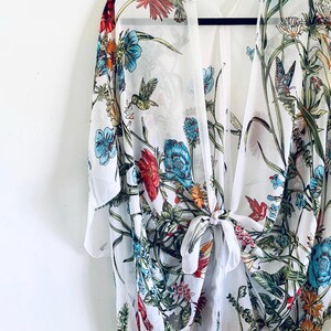 May include: A white kimono-style cover-up with a vibrant floral and hummingbird print. The design features red, blue, and green flowers with detailed hummingbird illustrations. The sheer fabric is tied at the waist, creating a flowing silhouette.