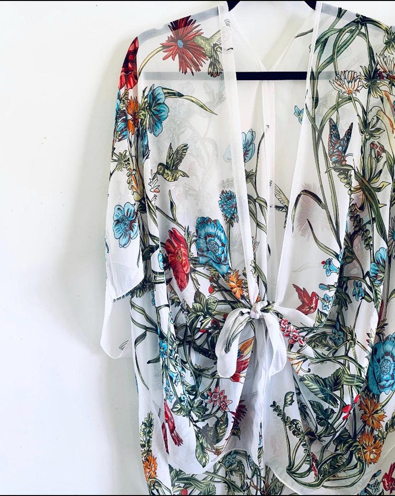 May include: A white kimono-style cover-up with a vibrant floral and hummingbird print. The design features red, blue, and green flowers with detailed hummingbird illustrations. The sheer fabric is tied at the waist, creating a flowing silhouette.