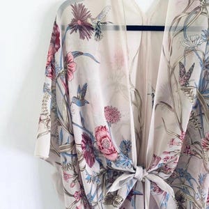 May include: A sheer, cream-colored kimono with a floral and hummingbird print. The design features pink, blue, and purple flowers, along with butterflies. The kimono is tied at the waist with a matching fabric tie and hangs on a black hanger.