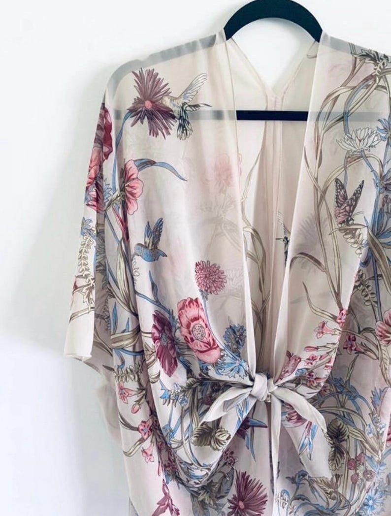 May include: A sheer, cream-colored kimono with a floral and hummingbird print. The design features pink, blue, and purple flowers, along with butterflies. The kimono is tied at the waist with a matching fabric tie and hangs on a black hanger.