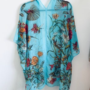 May include: A light blue, sheer kimono-style cover-up with a floral and hummingbird print. The design features various flowers in shades of red, orange, yellow, and white, with green leaves and stems. The garment is hanging on a black hanger.