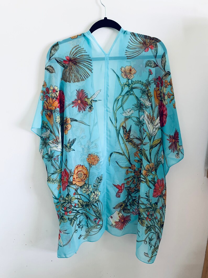 May include: A light blue, sheer kimono-style cover-up with a floral and hummingbird print. The design features various flowers in shades of red, orange, yellow, and white, with green leaves and stems. The garment is hanging on a black hanger.