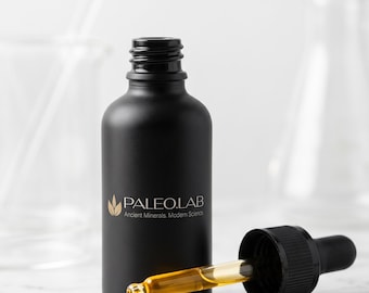 Pode incluir: Um frasco conta-gotas de vidro preto fosco com o texto PALEO.LAB Ancient Minerals, Modern Science impresso em branco. Uma pipeta de vidro com tampa preta repousa ao lado, com uma gota de líquido dourado visível sob o frasco.
