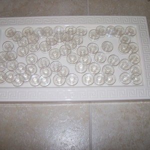 May include: A white ceramic tray with a Greek key border design, filled with a large quantity of clear plastic bobbins.