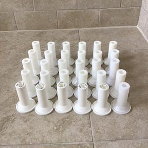 May include: A collection of twenty-four white plastic thread spools arranged in a grid. Each spool features a cylindrical body with a wide circular base and a hollow center, designed for holding sewing thread or similar craft materials.