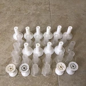 May include: An assortment of white and clear plastic spools and posts are arranged on a tiled surface. The spools have a circular design, while the posts have a cylindrical shape with a wide base. The items are likely for crafts or sewing.