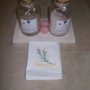 May include: Two glass jars with cork lids filled with bath products. The jar on the left is filled with rose bath foam and the jar on the right is filled with rose bath salts. Both jars have a label with the text "Rose Bath Foam" and "Rose Bath Salts". There are two pink bath bombs on a white tray with a decorative border. A white towel with the text "Chamomile" embroidered on it is on the counter.