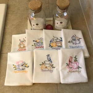 May include: Set of seven embroidered tea towels with a different design for each day of the week. Each towel features a bird, a teapot, and a floral design. The towels are white with colorful embroidery. The days of the week are embroidered on each towel: Monday, Tuesday, Wednesday, Thursday, Friday, Saturday, and Sunday.