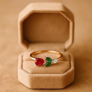 Può includere: Un anello in oro con una pietra preziosa rossa a forma di pera, due pietre verdi taglio marquise e una piccola pietra bianca rotonda. L'anello è presentato all'interno di un portagioie ottagonale color sabbia con interno in velluto.