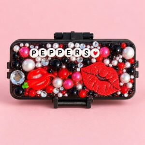 May include: A rectangular black case decorated with white, red, and black beads, rhinestones, and pearls. It features a red chili pepper charm, a red glittery lip-shaped charm, and white letter beads spelling PEPPERS with a red heart.