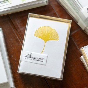 May include: A stack of white and gold stationery cards with a yellow ginkgo leaf design. The cards are packaged in a clear plastic box with the brand name "Ornament Paper & Letterpress" printed on the front.