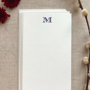 May include: A stack of white notepaper with a navy blue letter "M" printed at the top. The notepaper is on a neutral-colored surface, with red flowers and a branch with white buds in the background.