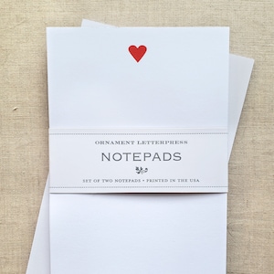 May include: Two white notepads with a red heart on the top notepad. The notepads are packaged together and have a label that reads "ORNAMENT LETTERPRESS NOTEPADS SET OF TWO NOTEPADS - PRINTED IN THE USA"