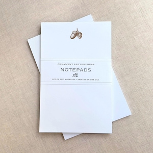May include: White notepads with a brown acorn illustration and the text "Ornament Letterpress" and "Notepads". The notepads are banded together with the text "Set of two notepads - Printed in the USA".